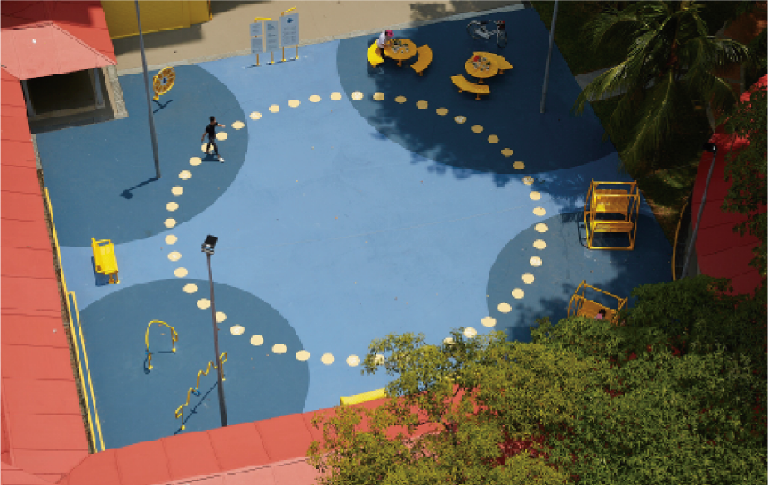 Aerial view of a blue outdoor community space with yellow play structures and benches, surrounded by trees and buildings.